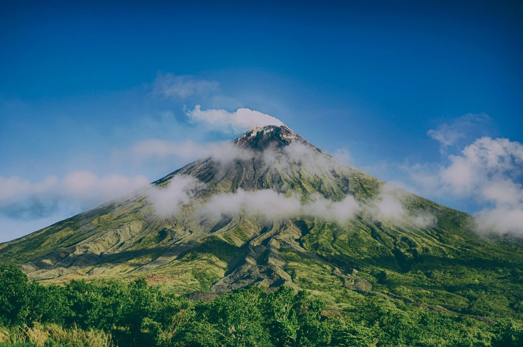 Logo de Tetis Recherche Ingénierie et Territoires superposé sur un grand volcan verdoyant entouré de nuages sous un ciel bleu